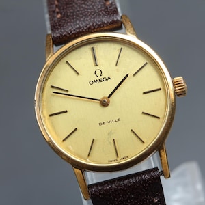 May include: An Omega De Ville wristwatch with a round gold-tone case and a matching gold-tone dial featuring thin black hour markers and hands. The watch includes a dark brown textured leather strap and displays the Omega logo and text.