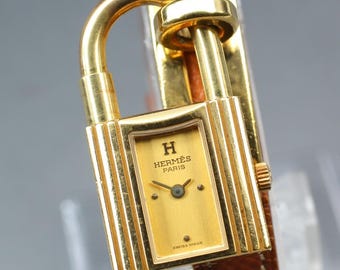 May include: A gold-toned wristwatch shaped like a padlock with a rectangular face, featuring the text HERMES PARIS and SWISS MADE on a gold dial. The watch has a metallic gold finish and is attached to a textured brown leather strap.
