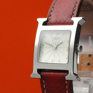 May include: A silver-toned wristwatch featuring a square case shaped like the letter H, a white dial with radiating lines, black Arabic numerals, and the text HERMES PARIS. The watch is attached to a textured dark red leather strap with stitching.