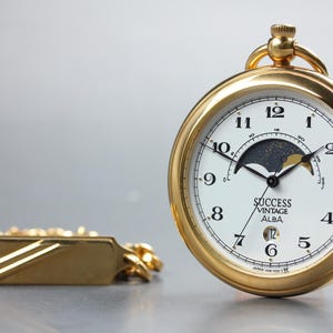 May include: A gold-tone pocket watch with a white dial, black Arabic numerals, and a moon phase display. The watch face features the text SUCCESS VINTAGE ALBA. A gold-tone chain with a rectangular bar attachment is positioned to the left of the watch.