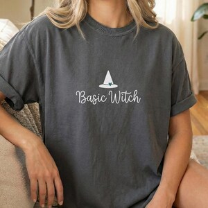 Può includere: Una t-shirt a maniche corte color grigio antracite con una grafica bianca di un cappello da strega sopra la scritta "Basic Witch" in un carattere corsivo bianco. La maglietta presenta un girocollo, una vestibilità ampia e maniche risvoltate.
