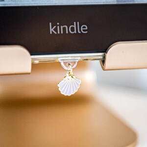 May include: A gold-colored metal stand holds a black Kindle e-reader. A small, light purple seashell charm with a gold-tone metal border hangs from a clear adhesive hook attached to the bottom center of the Kindle's frame.