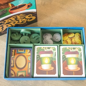 May include: A board game titled "Cafe Barbs" with various game pieces and cards. The game box is visible, along with a blue box containing green game pieces, gray stones, and cards featuring coffee cup illustrations.