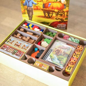 May include: A board game box with a yellow interior, filled with game pieces. The pieces include wooden blocks, cards, dice, and colorful tokens. The box has a lid with a picture of a farmer and pumpkins.