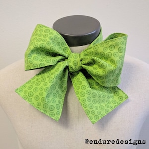 May include: A large green bow tie with a white and green circular pattern. The bow tie is tied and displayed on a mannequin.