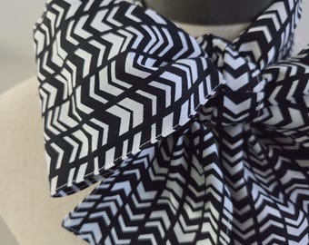 May include: A neck scarf featuring a bold black and white chevron pattern. The fabric is tied into a large, prominent bow at the front, creating a structured and layered appearance. The geometric arrow design repeats consistently across the entire surface.