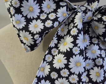 May include: A neck scarf featuring a repeating pattern of white daisy flowers with yellow centers on a solid black background. The fabric is tied in a knot, creating a bow-like appearance with two long, pointed ends draped over the chest area.