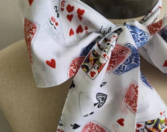 May include: A white fabric neck scarf featuring a repeating pattern of playing cards, including hearts, spades, and face cards in red, blue, and black ink. The scarf is tied in a knot, displaying various card suits and values across the surface.