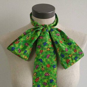 May include: A green bow tie with a pattern of colorful ladybugs. The bow tie is tied and displayed on a white mannequin.