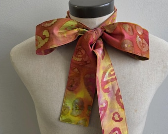May include: A neck scarf featuring a vibrant tie-dye pattern in shades of orange, yellow, and magenta. The fabric is tied into a bow at the center, with two long, rectangular ends hanging down. The scarf has a smooth texture and finished edges.