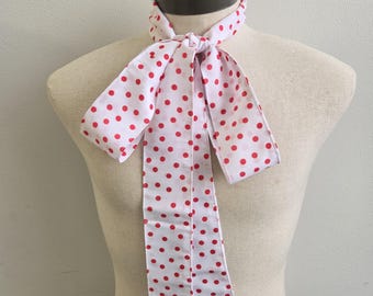 May include: A long, narrow neck scarf featuring a white background with a repeating pattern of small, vibrant red polka dots. The scarf is tied in a simple knot at the center, with two long, rectangular ends hanging down over the chest area.
