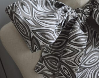 May include: A neck scarf featuring a repeating pattern of abstract, layered leaf-like shapes in shades of charcoal gray, silver, and white. The fabric is tied in a large, prominent bow around the neck of a beige mannequin torso.