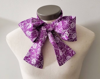 May include: A purple neck scarf featuring a white line-art floral pattern. The scarf is tied in a large, prominent bow at the center, with two long, wide fabric tails extending downward. The fabric has a smooth texture and a vibrant purple hue.