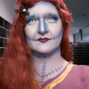 May include: A woman with red hair and blue makeup is dressed as a character from the film 'The Nightmare Before Christmas'. She has stitches drawn on her face and neck, and is wearing a brown and purple dress.