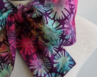 May include: A neck scarf tied in a bow, featuring a deep purple background with a repeating pattern of circular, sunburst-like shapes in vibrant shades of pink, turquoise, yellow, and light blue, displayed on a mannequin neck.