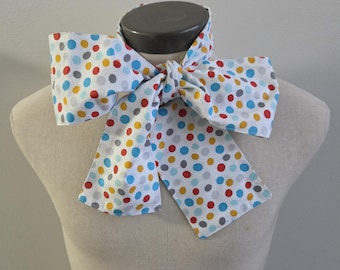 May include: A white fabric neck scarf featuring a repeating pattern of multicolored polka dots in shades of red, blue, yellow, and grey. The scarf is tied in a large, prominent bow at the center, with two long, wide ends hanging down from the knot.