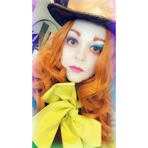 May include: A woman wearing a costume with a large, brown top hat, a yellow bow tie, and bright orange curly hair. She has colorful makeup on her face, including blue and pink eyeshadow.