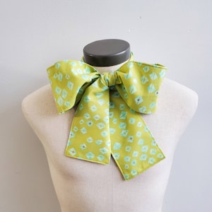 May include: A green bow tie with a light blue geometric pattern. The bow tie is tied and displayed on a white mannequin.