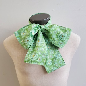 May include: A green bow tie with a white and gold circular pattern. The bow tie is tied and displayed on a mannequin.