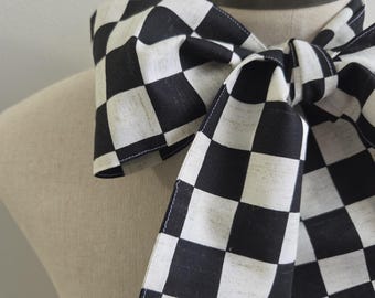 May include: A neck scarf featuring a black and white checkered pattern. The fabric is tied in a bow around the neck of a mannequin. The scarf has visible stitching along the edges and a repeating square grid design throughout the material.