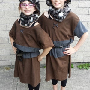 May include: Two children wearing brown hooded tunics with fur trim and black leggings. They are wearing black shoes and have matching fur hats.