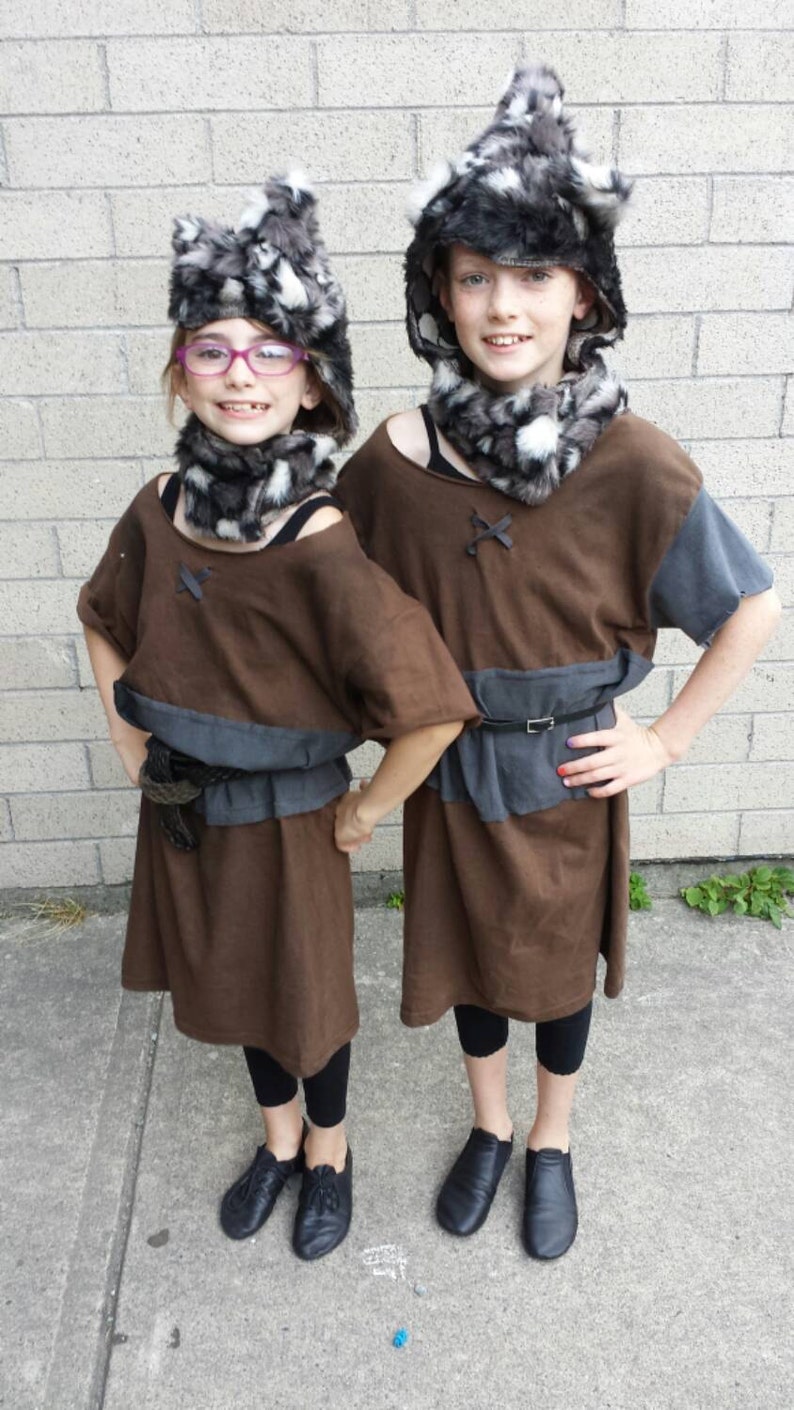 May include: Two children wearing brown hooded tunics with fur trim and black leggings. They are wearing black shoes and have matching fur hats.