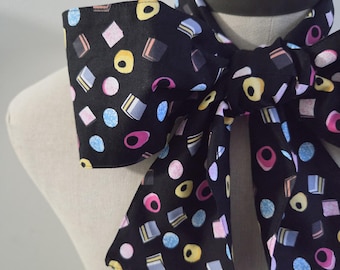 May include: A black neck scarf featuring a repeating pattern of colorful candy shapes including yellow rings, pink squares, blue textured circles, and layered rectangular sweets. The fabric is tied in a large bow around a neutral-toned mannequin neck.