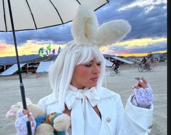 May include: A woman wearing a white coat and a white bow tie, with white bunny ears and a white wig, holds a small stuffed animal and a white umbrella. She is standing in a desert setting with a sunset in the background.