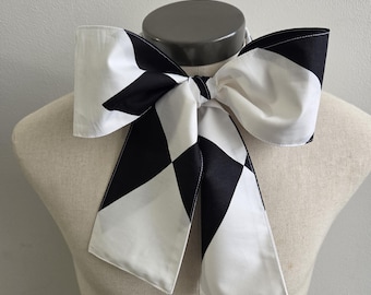 May include: A large fabric bow tie featuring a bold geometric pattern of alternating black and white diamond shapes. The accessory is tied in a structured knot with two long, wide tails hanging down, showcasing a crisp, clean contrast.