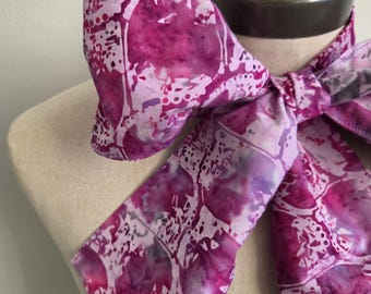 May include: A neck scarf featuring a vibrant magenta and white batik-style pattern with abstract circular and geometric shapes. The fabric is tied into a large bow around the neck, displaying a smooth texture and a repeating, intricate design throughout.