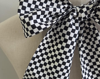 May include: A large fabric bow tie featuring a black and white checkered pattern with hand-painted style squares. The accessory is tied in a knot and drapes down with two long, wide tails, showcasing a repeating geometric grid design throughout.