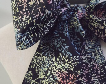 May include: A black fabric neck scarf featuring a repeating pattern of fern-like leaves in shades of pastel pink, blue, yellow, and white. The scarf is tied in a large bow at the neck, with two long, wide ends hanging down, displaying the intricate botanical print.