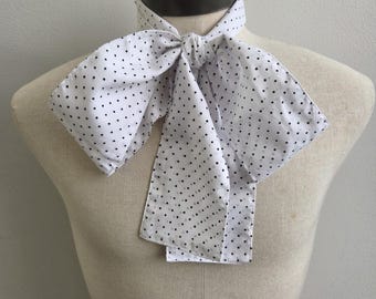 May include: A white neck scarf featuring a repeating pattern of small black polka dots. The scarf is tied in a large, prominent bow at the center, with two wide, rectangular ends hanging down from the knot, resting against the chest area.