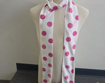 May include: A long, narrow white scarf featuring a repeating pattern of vibrant pink polka dots. The scarf is draped around the neck of a mannequin, displaying a smooth fabric texture with evenly spaced circular motifs in a bright magenta hue.
