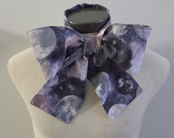 May include: A neck scarf tied in a large bow featuring a dark purple and grey space-themed print with multiple moon phases and celestial patterns. The fabric has a smooth texture and is wrapped around the neck of a neutral-toned display mannequin.