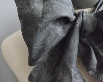 May include: A dark gray fabric neck accessory tied in a large bow. The material features a subtle, mottled, or marbled pattern throughout, with visible stitching along the edges of the fabric loops and tails, creating a structured appearance.