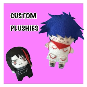 May include: Two handmade felt plushies against a bright pink background. One plushie has black hair with a red streak and a black outfit with the text CCCP. The other has blue hair, a white outfit with red accents, and a red scarf. Text reads CUSTOM PLUSHIES.