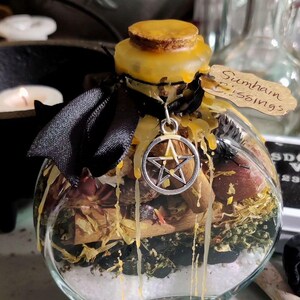 May include: A clear glass jar filled with dried herbs, white granules, and black stones. The jar is sealed with a cork and yellow wax, adorned with a black ribbon and a silver pentacle charm. A small tag reads "Sumhain Blessings".