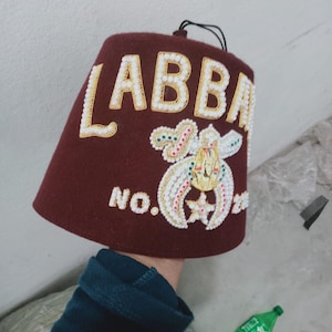 Op de afbeelding: Een bordeauxrode fez-hoed met goud- en wit geborduurde letters en versieringen. De hoed heeft het woord "LABBA" en het nummer "22" met een decoratief embleem. Een zwart touwtje is aan de bovenkant van de hoed bevestigd.