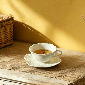 May include: A white teacup and saucer set filled with tea, placed on a burlap runner atop a white cabinet. A wicker basket and dried flowers are in the background. The teacup has a scalloped edge and a delicate handle.
