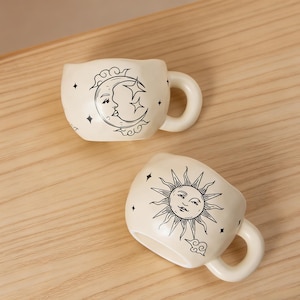 May include: Two off-white ceramic mugs with black celestial designs. One mug features a crescent moon and stars, the other a smiling sun. Both have rounded handles and are set on a wooden surface.