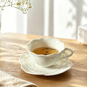 May include: A white teacup and saucer set with a floral design, filled with tea. The cup has a delicate handle and sits on a matching saucer. The set is on a wooden table, with a book and a small plate nearby.