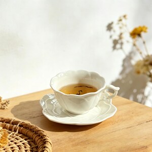 May include: A white teacup and saucer set filled with tea, resting on a wooden table. The cup and saucer have a scalloped edge design. Dried flowers and a woven basket are also visible in the image.