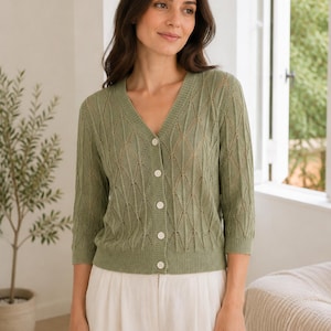 May include: A sage green V-neck cardigan featuring a delicate openwork diamond knit pattern, three-quarter length sleeves, and a front button closure with five small white buttons. The garment has a ribbed neckline, cuffs, and hem.
