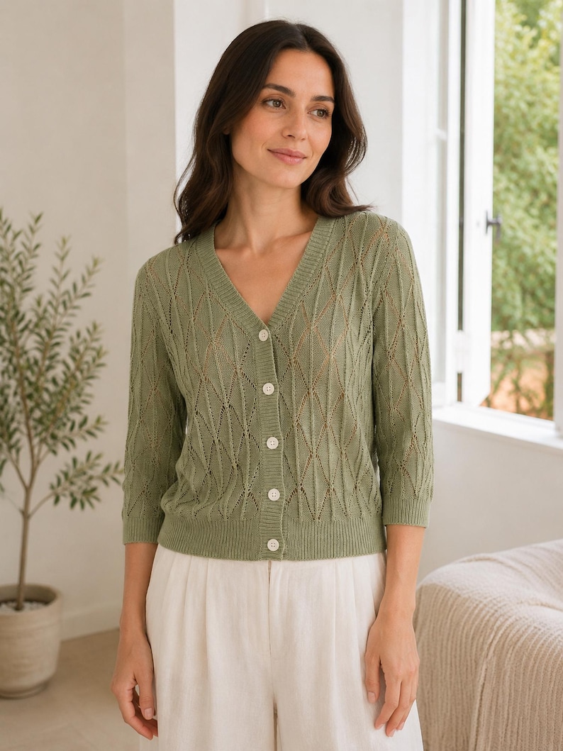 May include: A sage green V-neck cardigan featuring a delicate openwork diamond knit pattern, three-quarter length sleeves, and a front button closure with five small white buttons. The garment has a ribbed neckline, cuffs, and hem.