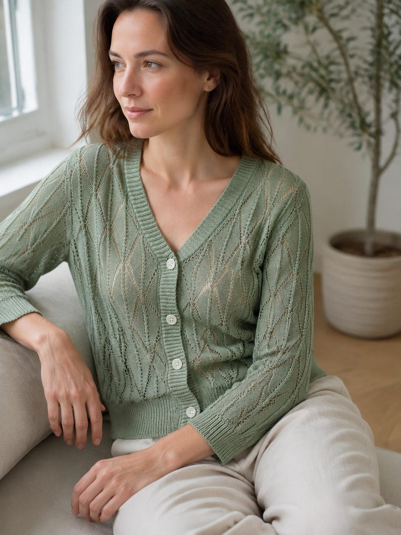 May include: A sage green V-neck cardigan featuring a delicate openwork knit pattern with vertical diamond-shaped motifs. The garment has long sleeves, a ribbed hem and cuffs, and is fastened down the front with four small, round, light-colored buttons.
