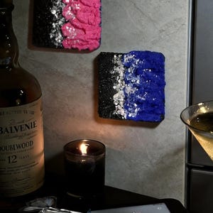 May include: Two square textured wall art pieces featuring a black base, a central strip of silver glitter, and a vibrant colour block of either hot pink or royal blue. The art is displayed alongside a bottle of Balvenie Doublewood 12 whisky.