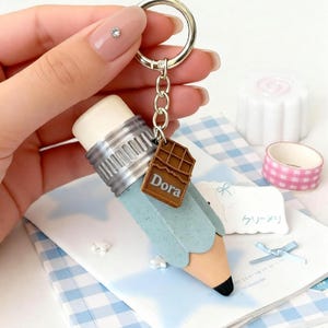 Op de afbeelding: Een potloodvormige sleutelhanger met een lichtblauwe behuizing, een beige punt met een zwarte stift, een zilverkleurige metalen huls en een witte gum. Een kleine bruine chocoladereep-bedel met de naam Dora is aan de ring bevestigd.