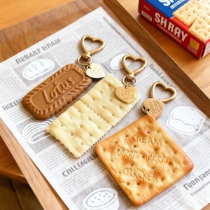 Puede incluir: Tres llaveros con forma de galleta y cierres dorados en forma de corazón. Uno es una galleta Lotus rectangular marrón, otro es una galleta salada rectangular pálida y el tercero es una galleta salada cuadrada dorada con el texto CREAM CRACKER.