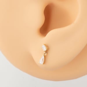 May include: Close-up of two earrings on a mannequin ear. One earring is gold-toned with a teardrop-shaped charm, and the other is silver-toned with a similar charm. Both earrings have a small, square-shaped gem at the top.